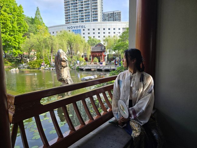 Wing staring at the pond within a Chinese garden. She is wearing neo-Chinese styled clothing.