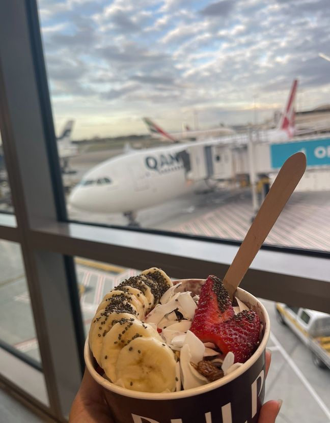 Photo of Wing holding an açai bowl in front of the airport window.